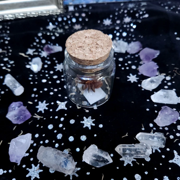 A small corked jar holding herbs, surrounded by amethyst and clear quartz on a dark starry cloth