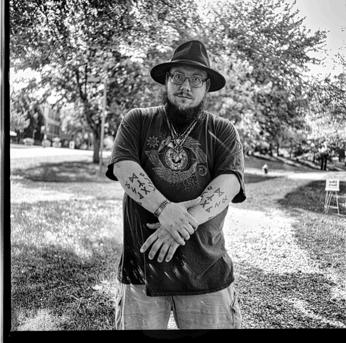 Sarenth Óðinsson, a bearded man in a wide-brimmed hat and a t-shirt with a Norse raven design, standing in a park with Elder Futhark runes tattooed along both forearms.