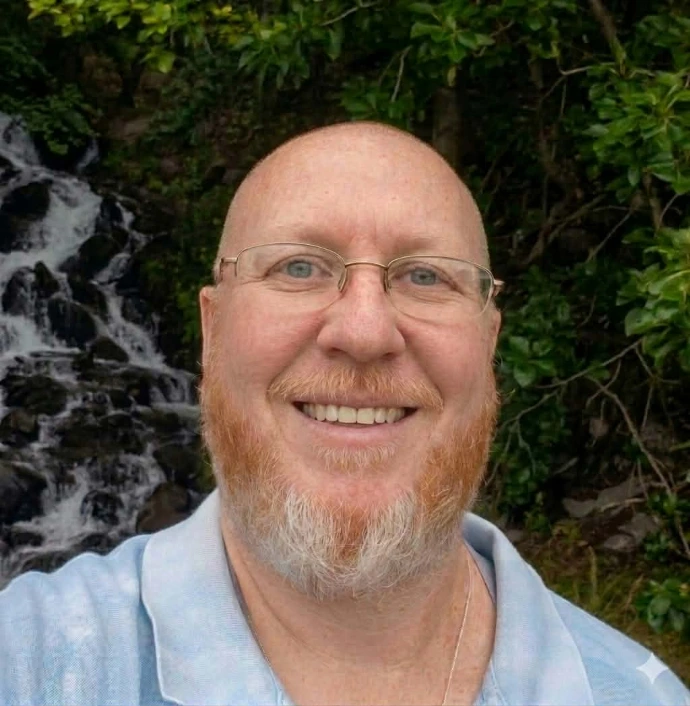 Milo Kincaid, a smiling man with a red and white beard and wire-rimmed glasses in a light blue polo shirt, standing in front of a waterfall.