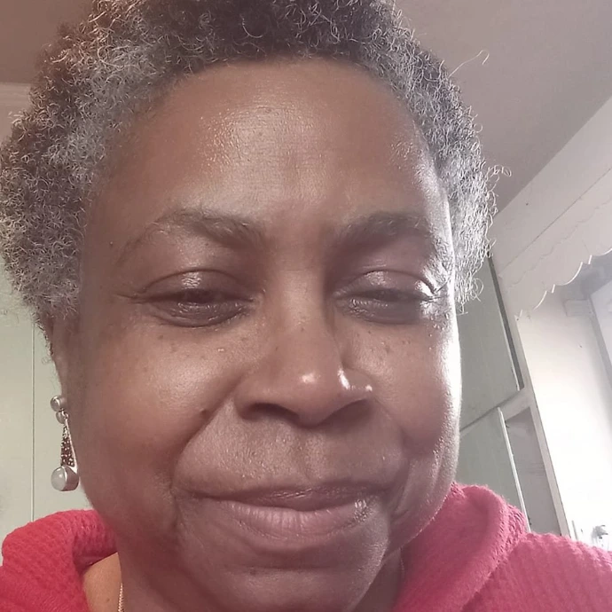 A close-up selfie of a woman with short, curly grey hair, wearing a red hooded sweatshirt and dangling metallic earrings, smiling gently at the camera.   