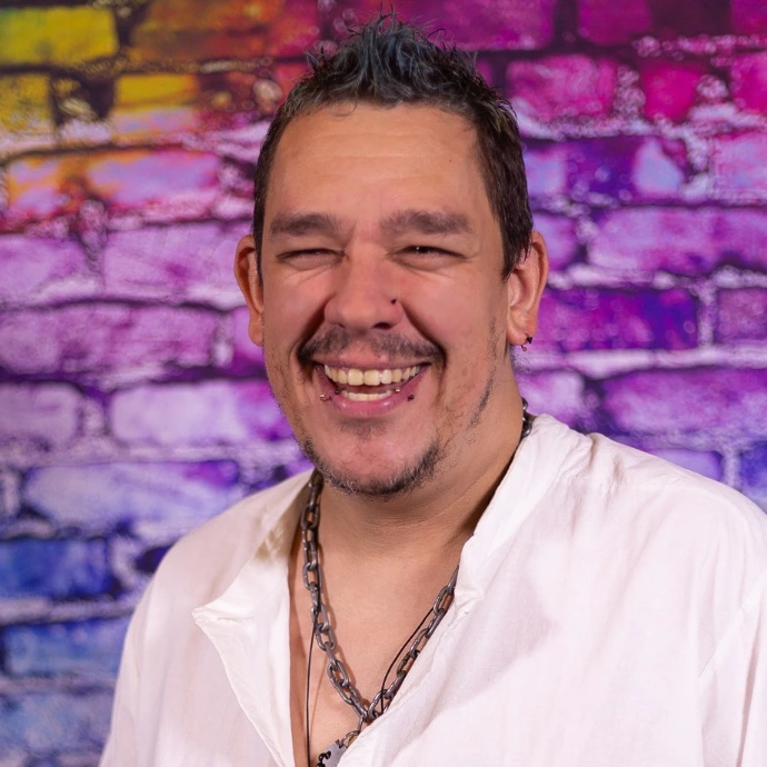 A headshot of a smiling man with short spiked hair, facial piercings, and a silver chain necklace, wearing a white shirt against a colorful brick wall backdrop.   
